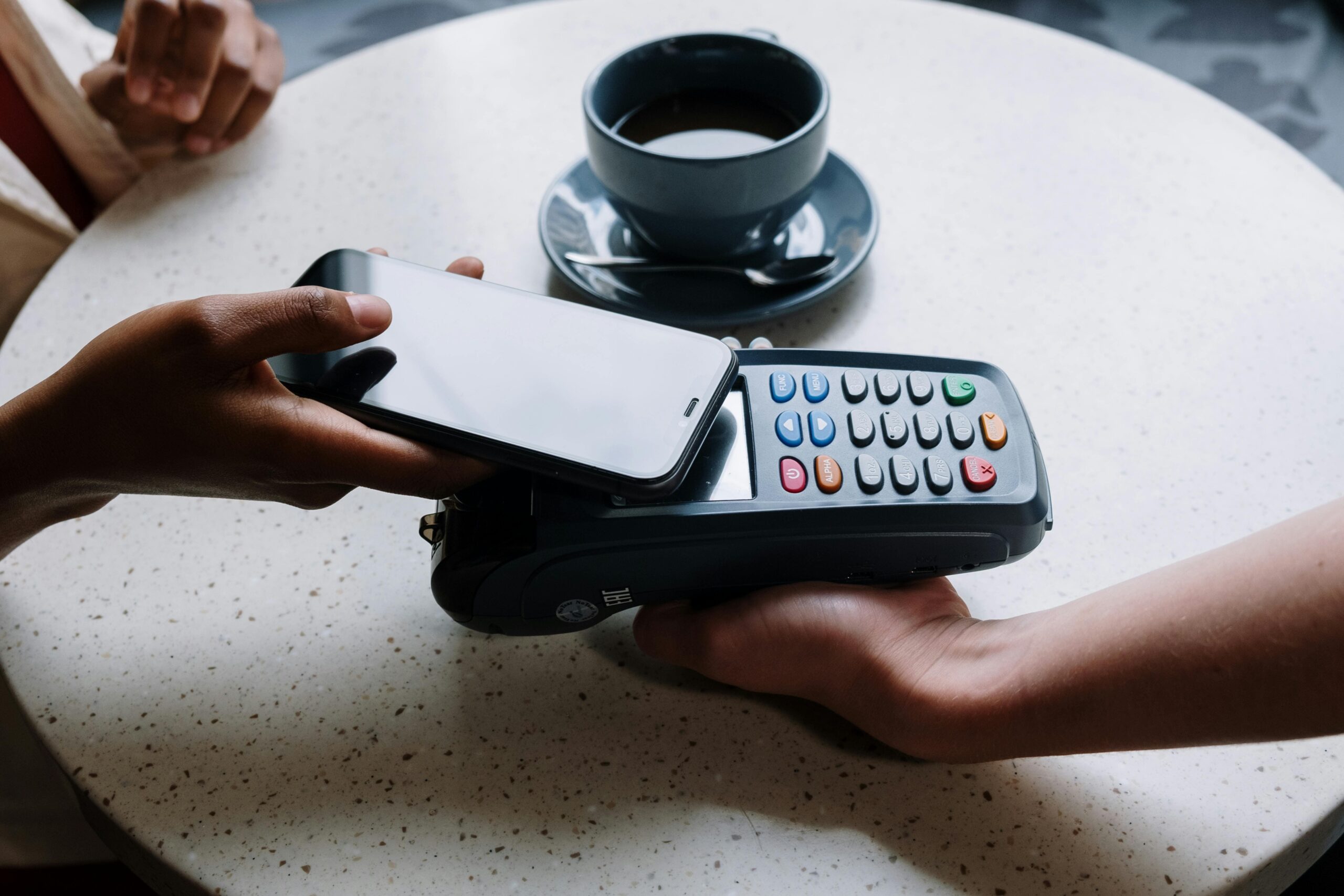 A close-up of two hands; one hand is holding a smartphone over a payment terminal, and the other hand is holding the terminal. A cup of coffee is visible in the blurred background.
