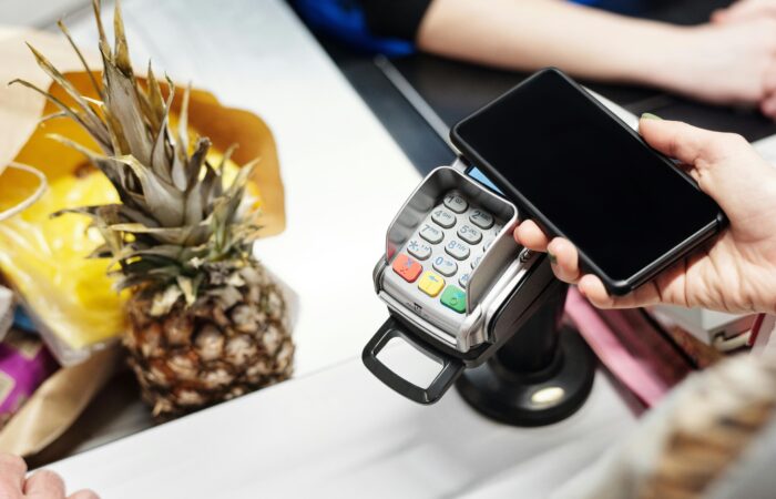 A woman is making a payment with her smartphone at a store checkout, holding the phone over a card reader. A pineapple and a paper bag are visible on the counter.