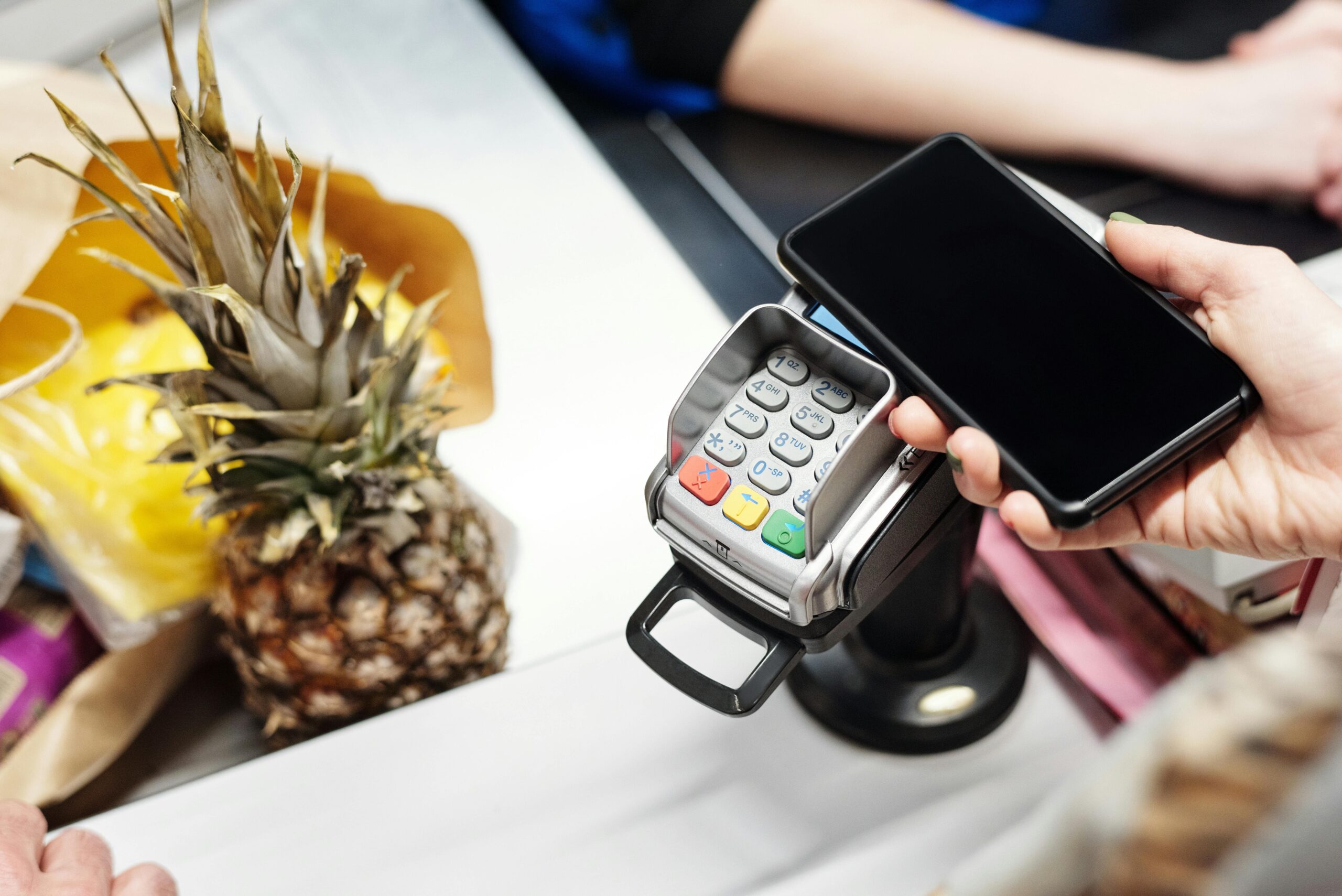 A woman is making a payment with her smartphone at a store checkout, holding the phone over a card reader. A pineapple and a paper bag are visible on the counter.