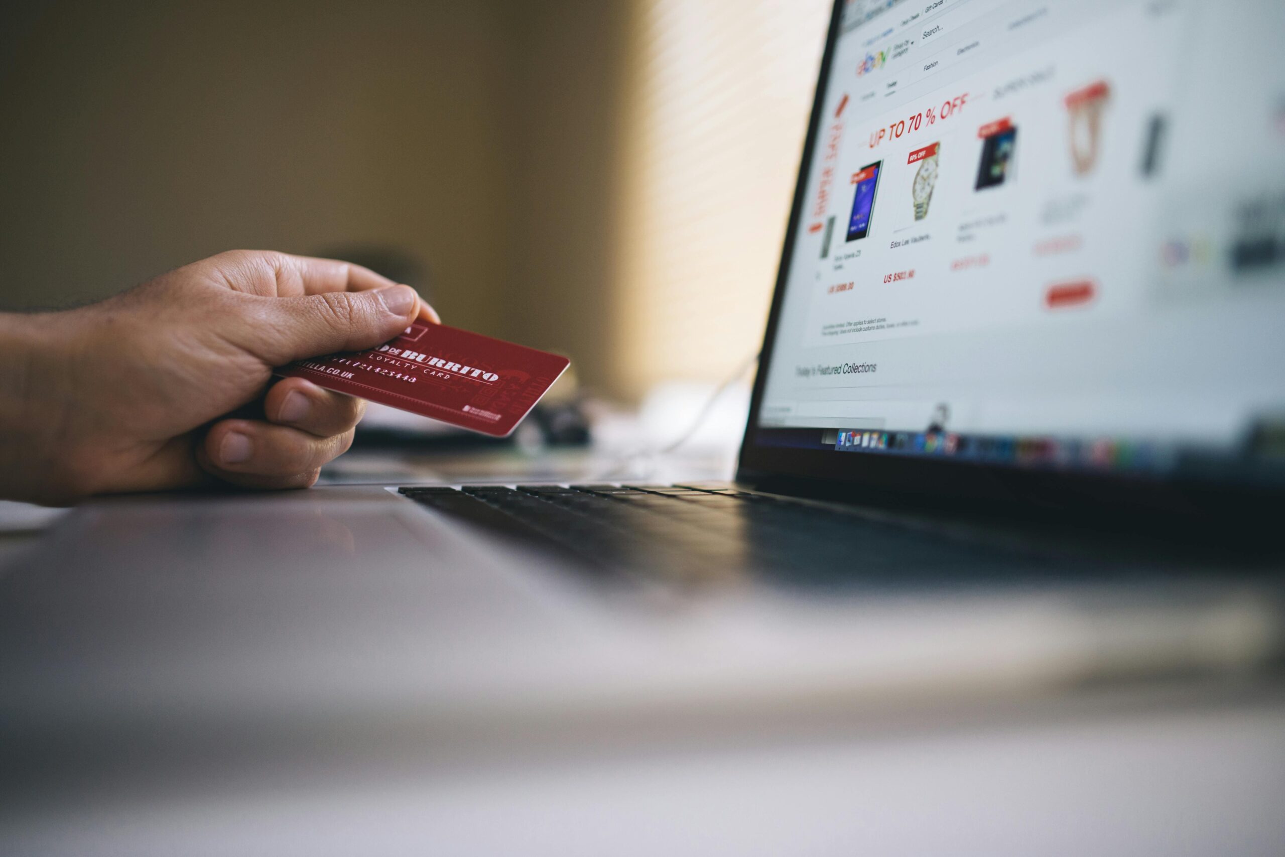 A hand holds a red credit card in front of a laptop screen displaying an online shopping website with various products.